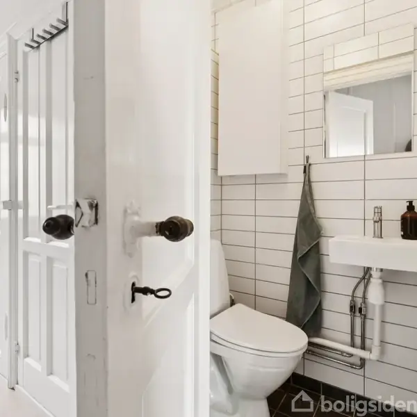 Et lille badeværelse med hvid flisevæg, toilet og håndvask. Et mørkegrønt håndklæde hænger ved siden af vasken. En dør åbner ind til et lyst rum.