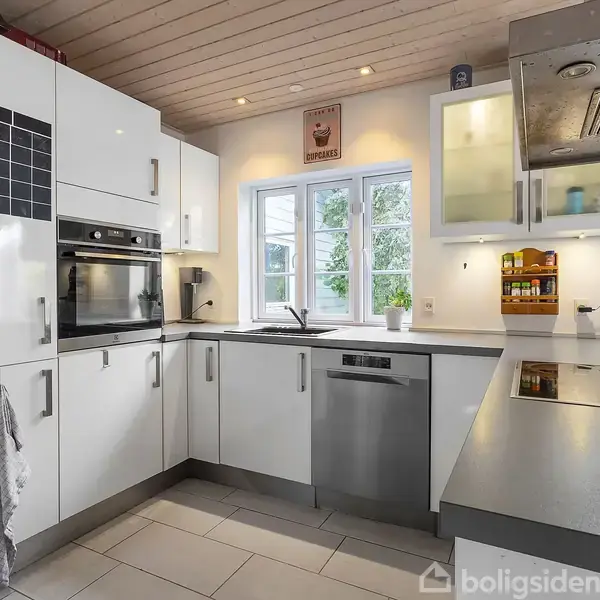 Hvidt køkken med indbyggede apparater. En ovn og opvaskemaskine er synlige. Store vinduer i baggrunden giver udsigt til grøn natur. Tekst: "This Month" på et magnetisk skema.