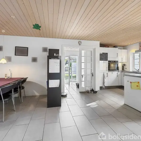 Køkkenet har lyse fliser og træloft. Et stort bord med lilla dug og stole står i det ene hjørne. Der er også køkkenskabe, hvidevarer og en halvvæg med emhætte.