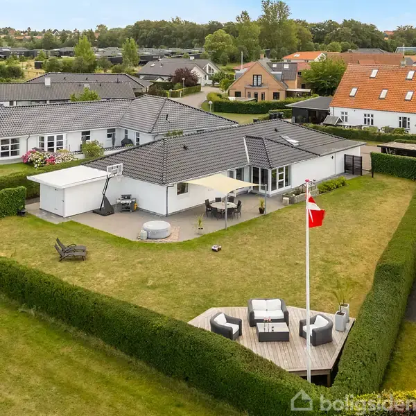 En hvid bolig med sort tag står på en flisebelagt grund, omkranset af hække. I baggrunden er der en sø og flere huse, omkranset af grønne områder.