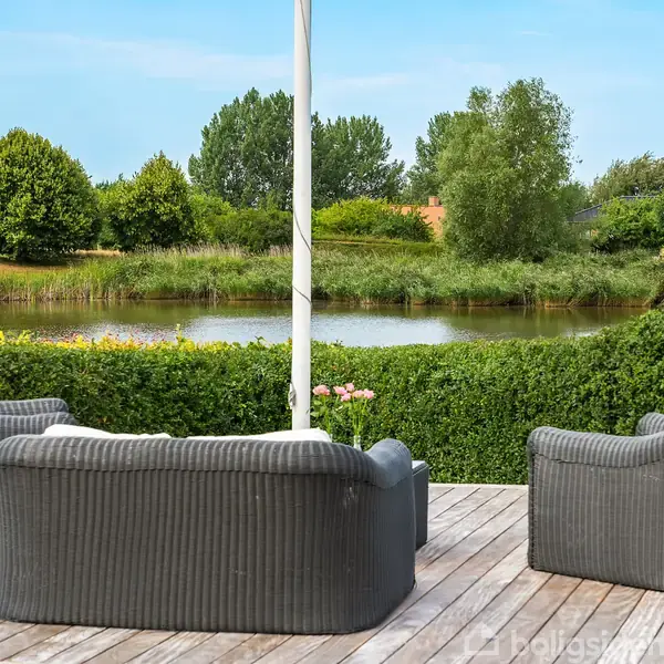Tre grå udendørs sofaer står på en træterrasse. De er rettet mod en sø omgivet af grønne buske og træer i en have.