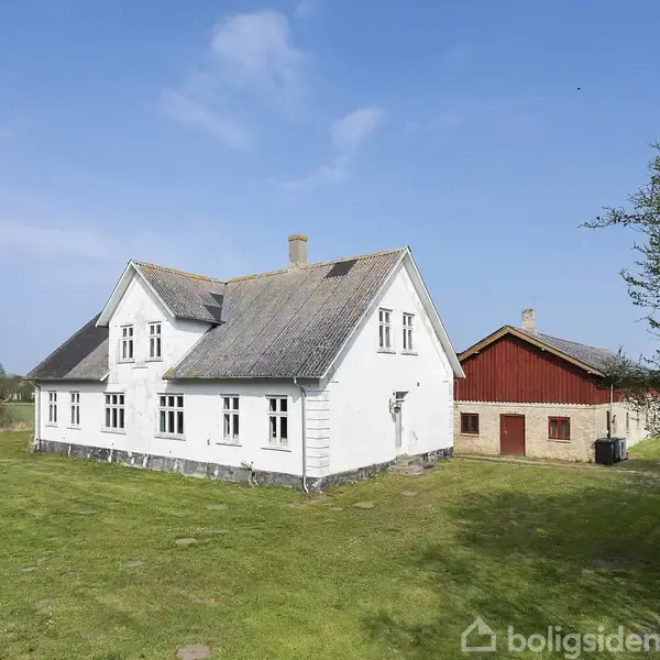 Hvidt, to-etagers hus står stille på en grøn græsplæne ved siden af en rød lade. Landskabet er åbent under en klar blå himmel.