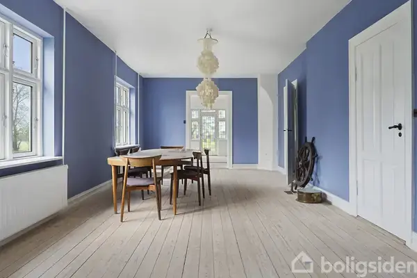 Et spisebord med stole står centralt i et langt rum med blå vægge og lyse trægulve. En stor lampe hænger over bordet. Vinduerne slipper dagslys ind.