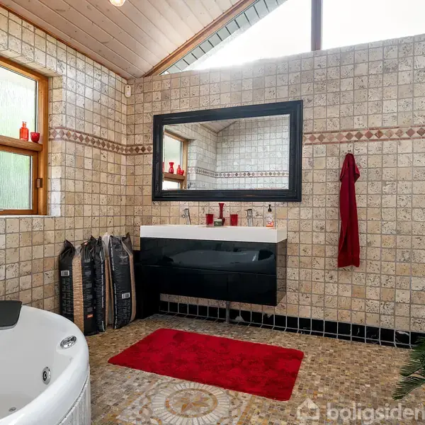 Boblebad står stille i et badeværelse med mosaikfliser på gulvet og væggene. Spejl, håndvask og rødt håndklæde skaber en moderne atmosfære. Røde stearinlys pryder vindueskarmen.