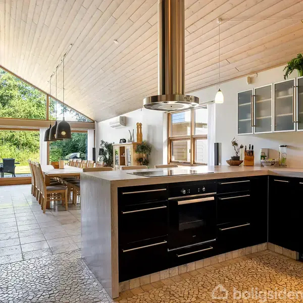 Køkken med sort køkkenbord, integreret ovn og emhætte, åbner op mod en spisestue med lang spisebord, store vinduer og udsigt til en træfyldt have.