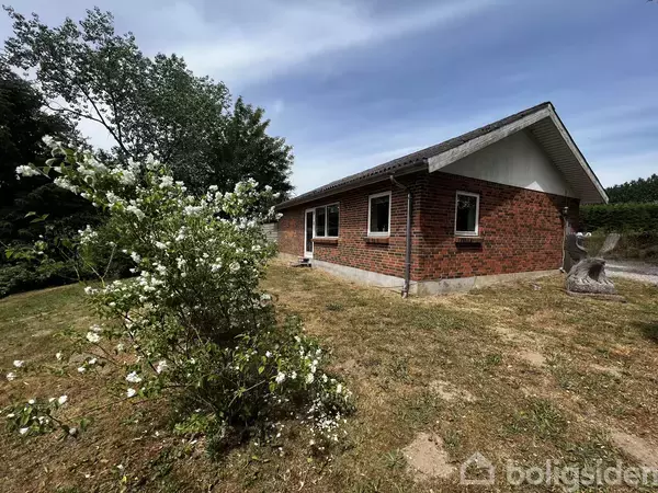 Et rødstenshus med gråt tag står på en tør græsplæne. Foran huset blomstrer en stor busk med hvide blomster. Træer omkranser området i baggrunden.