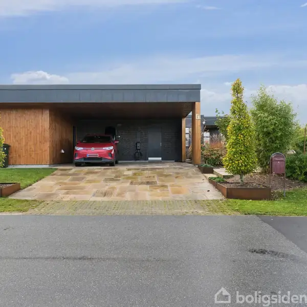 En moderne, fladtag træhus med en parkeret rød bil i carporten, omgivet af trimmet have med buske. Skydedøren til huset ses bag bilen.