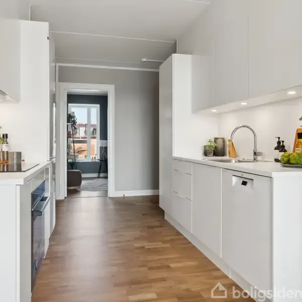 Et moderne køkken med hvide skabe og trælaminatgulv. Der er planter og køkkenredskaber på bordpladerne. Rummet leder ind til en stue med et vindue, der viser en balkon udenfor.