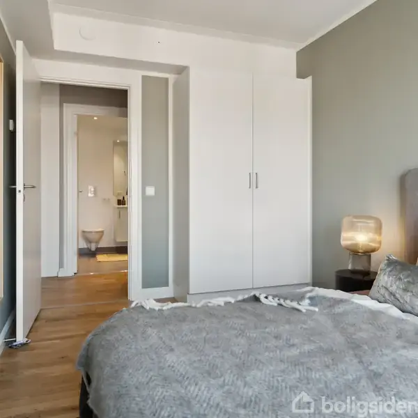 Soveværelse med gråt sengetæppe, pude og natbordslampe. Skabsdøre står på klem, og en dør åbner til et badeværelse med toilet. Trægulv og grå vægge præger rummet.