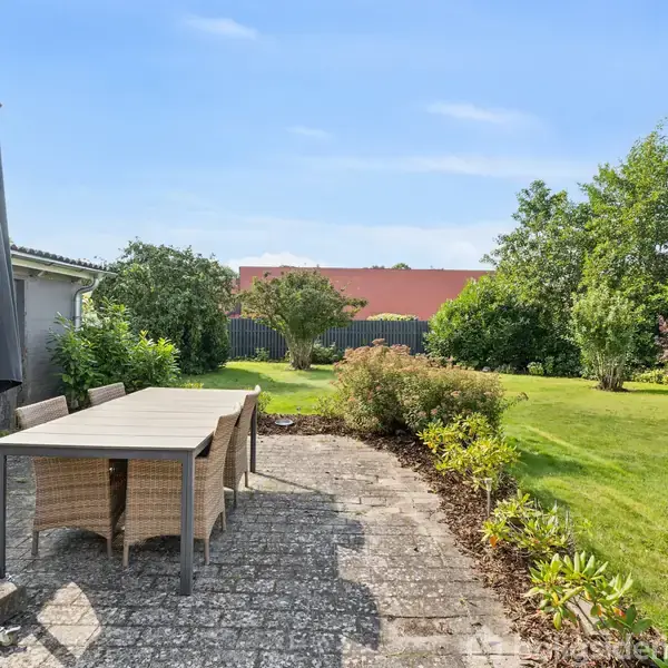 Et havesæt med bord og stole står på en flisebelagt terrasse. Foran er der en grøn græsplæne med buske og træer i baggrunden under en klar blå himmel.