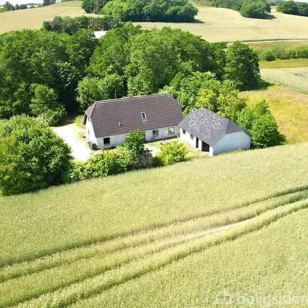 Et hus og en længe ligger omgivet af træer og marker med grønne afgrøder. Bygningerne er placeret i landlige omgivelser i et åbent, bakket landskab.