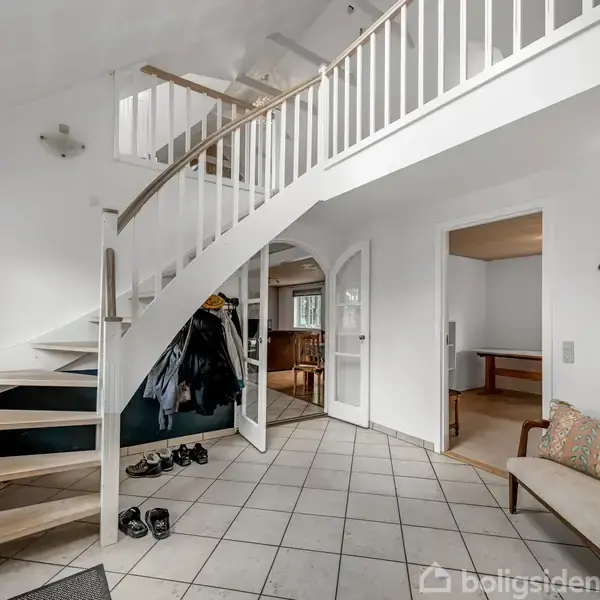 En åben entré med en hvid trappe, der fører til øverste etage. Væggene er hvide, og der er flisegulv. Skohylder og en bænk med farverig pude står langs væggen.