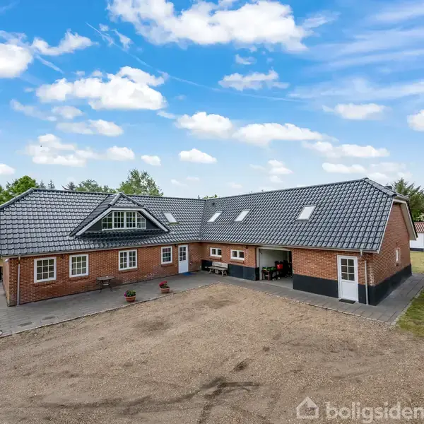 En stor, vinkelformet murstensbygning med gråt tegltag står omgivet af en grusvej og græsplæner i et landligt område under en blå himmel med hvide skyer.