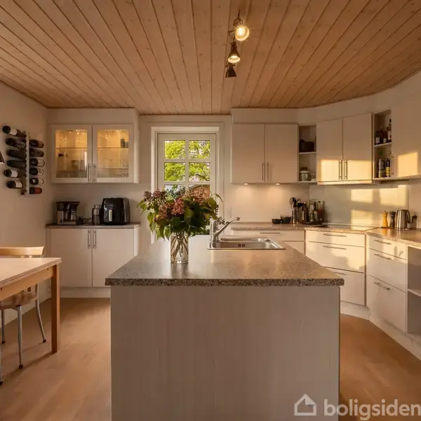 Et moderne køkken med lyst træloft og gulv. Køkkenøen har en vase med blomster. Til venstre står et spisebord ved vinduer, og højre side har skabsplads og køkkenmaskiner.