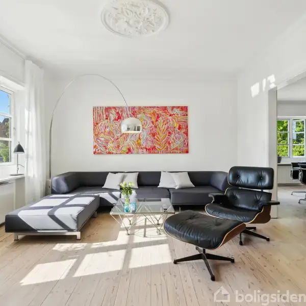En moderne stue med en sort lædersofa og lænestol, dekoreret med farverigt kunstværk. Store vinduer giver rummet lys. I baggrunden ses en spisestue med spisebord og stol.