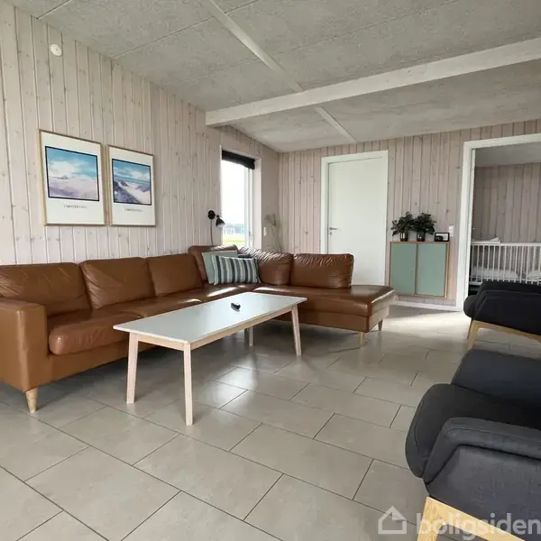 Brun lædersofa arrangeret i hjørne, flankeret af sort lænestol. Sofabord i træ med hvidt top. Trævægge, to billeder hænger, skaber hyggelig stue. Dør åbner til ekstra rum.