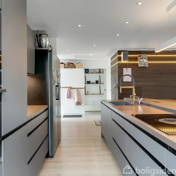 En moderne køkkenø med indbygget kogeplade og vask. Omgivet af grå skabe og trægulv. I baggrunden ses en spiseplads og åben reol. Ingen synlig tekst.