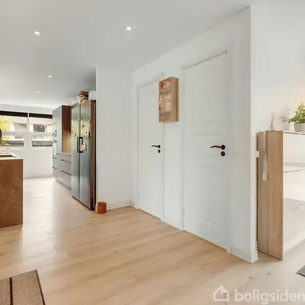 Køkken med træparketgulv og hvide vægge, indeholder moderne brune køkkenelementer, stor mørk grå køleskab, store vinduer samt hvide døre, der fører til andre rum, og glasmontre med planter.