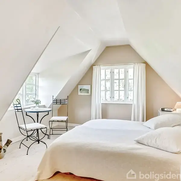 Et soveværelse med en stor seng under et skråt loft. Et rundt bord med to stole står ved et vindue. Rummet er lyst og indbydende.
