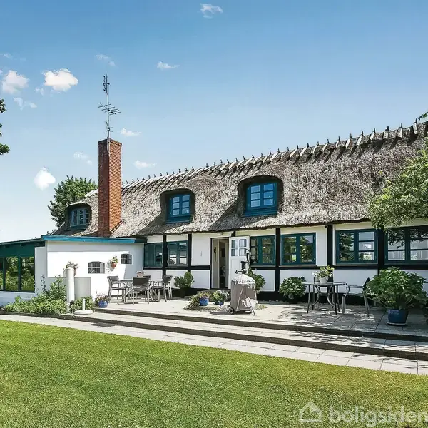 Stråtækt hus med hvid facade og blå vinduer står på en velplejet græsplæne. Der er en terrasse foran huset med møbler, og omgivelserne inkluderer træer og blå himmel.
