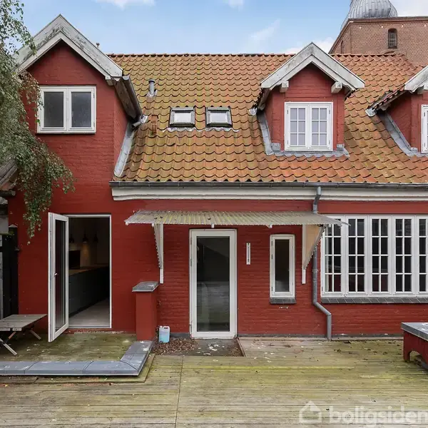 Rødt murstenshus med to etager og flere gavle, åbne døre til terrasse med trægulv, omgivet af sort hegn og delvist synligt træ i baggrunden.