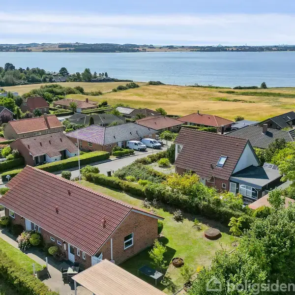 Rødt murstenshus med have ligger mellem andre huse i et grønt boligområde med udsigt over havet. Bygningen har et rødt tegltag og er omgivet af buske og træer.