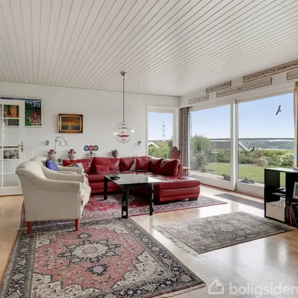 Stue med rød sofa og hvid lænestol står på orientalske tæpper. Store vinduer åbner op til en have med havudsigt. Reoler og lamper skaber en hyggelig atmosfære.