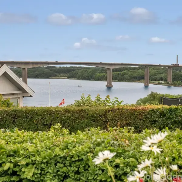 Bro strækker sig over en flod med skovklædte skråninger i baggrunden. Forgrund: En have med blomster og en halvtag, der leder øjnene mod vandet.