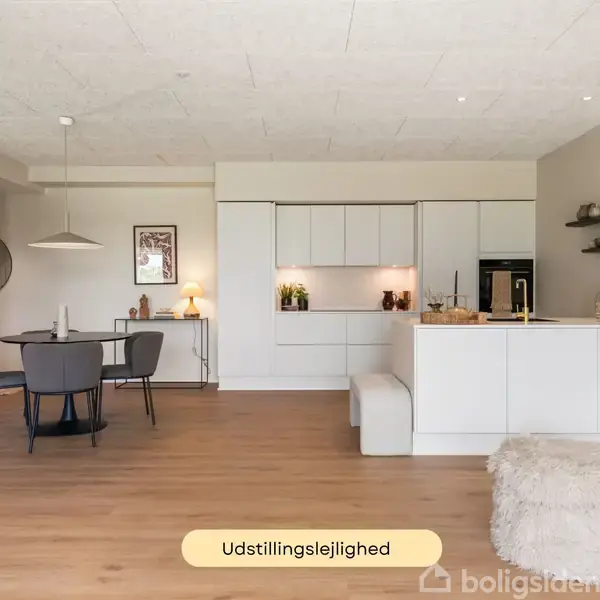 Køkken med hvidt interiør og ø i moderne lejlighed; en træbord med fire stole er placeret til venstre. Et skilt med teksten "Udstillingslejlighed" er synligt.