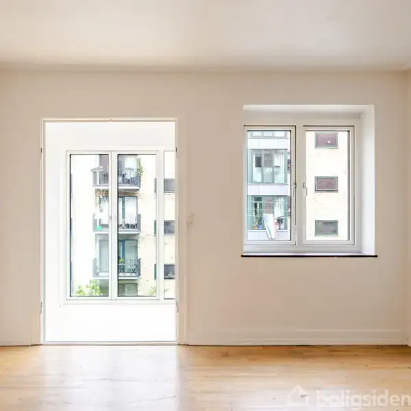Et tomt, lyst rum med trægulv; et åbent dørparti og et vindue vender ud mod en lysfyldt gård med bygninger. Adjacent er hvide vægge og lofter, der giver et rent look.