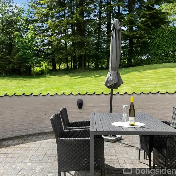 Rattanmøbler står omkring et bord med en vinflaske og glas på en fliseterrasse. En parasol er lukket. I baggrunden ses en velplejet græsplæne omgivet af grønne træer.