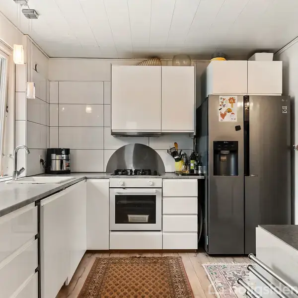 Et moderne køkken med hvide skabe og sorte bordplader. Køleskabet er af rustfrit stål, og over komfuret er et udsugningssystem. Et tæppe ligger på trægulvet.