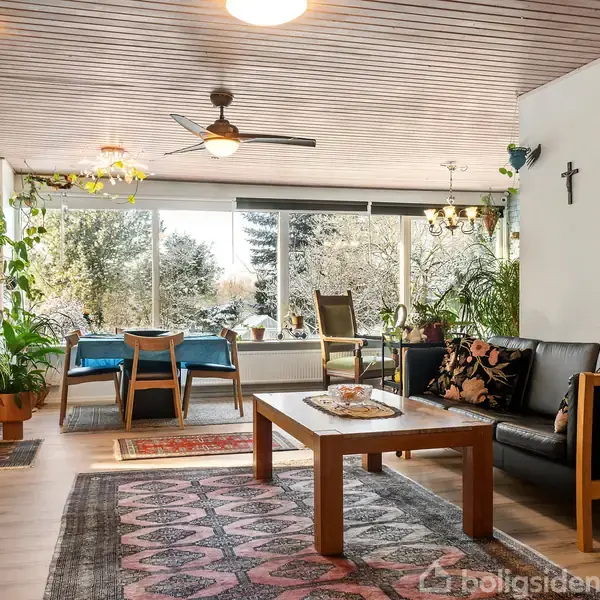 Stue med sofa og sofabord foran store vinduer. Planter pryder rummet. I baggrunden ses et spisebord med stole. Luften i rummet cirkuleres af en loftsventilator.