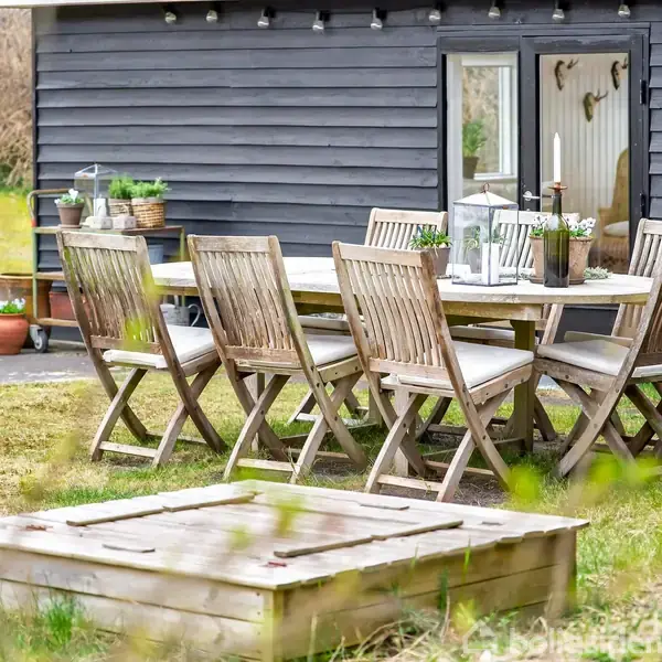 Træmøbler står ude; bord omgivet af otte stole. I baggrunden ses et sortmalet hus. Planter og lanterner pynter bordet, der står på en græsplæne med en trækasse foran.