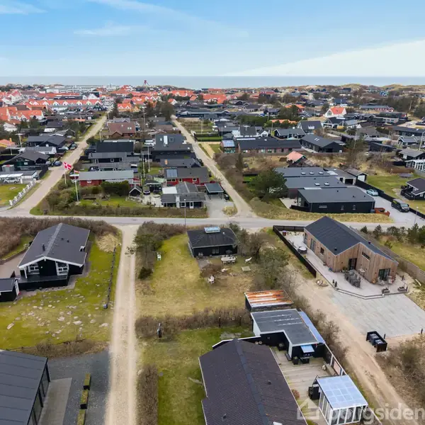 Huse ligger tæt ved grusveje i et villakvarter med grønne områder, mens røde tage i baggrunden indikerer nærhed til en by med flere bygninger og kystlinjen i horisonten.