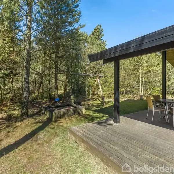 Terrasse med borde og stole står stille. Træterrassen er omkranset af skov og græsplæne. Et gyngestativ ses i baggrunden blandt træerne.