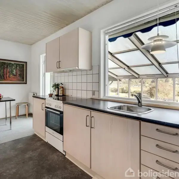 Et køkken med hvide skabe og sort bordplade. En håndvask og komfur er placeret under et stort vindue. En spisestue ses i baggrunden med maleri og ur på væggen.