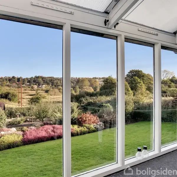 Ruder i en veranda viser en udsigt over en frodig have med græsplæne og blomstrende buske; i baggrunden ses et kuperet landskab med træer og buske.