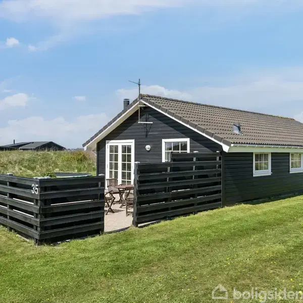 Sommerhus med sort træbeklædning står på græsplæne, omkranset af sort stakit med tegnet nummer 35. Solrig himmel i baggrunden giver et indtryk af en rolig, åben beliggenhed.