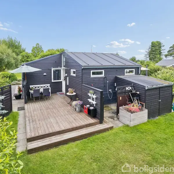Sort træhus står stille på en græsplæne med terrasse i solen. Omgivet af buske og træer, er der også havemøbler under en parasol.