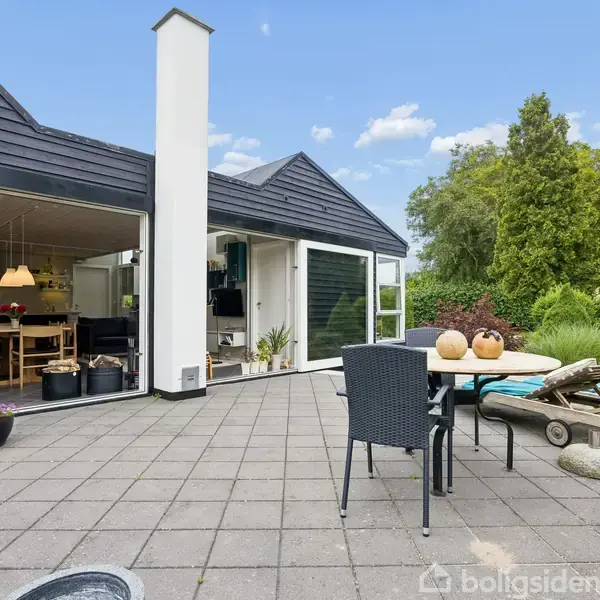 Moderne terrasse med sorte fliser og havemøbler, der inkluderer en stol og et bord. Åbne døre fører ind til en stilfuld spisestue. Omgivet af grønne træer og buske.