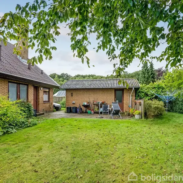 Hus set udefra med en terrasse med stole og bord, omgivet af græsplæne og grønne træer. En vaskesnor ses til højre i haven.