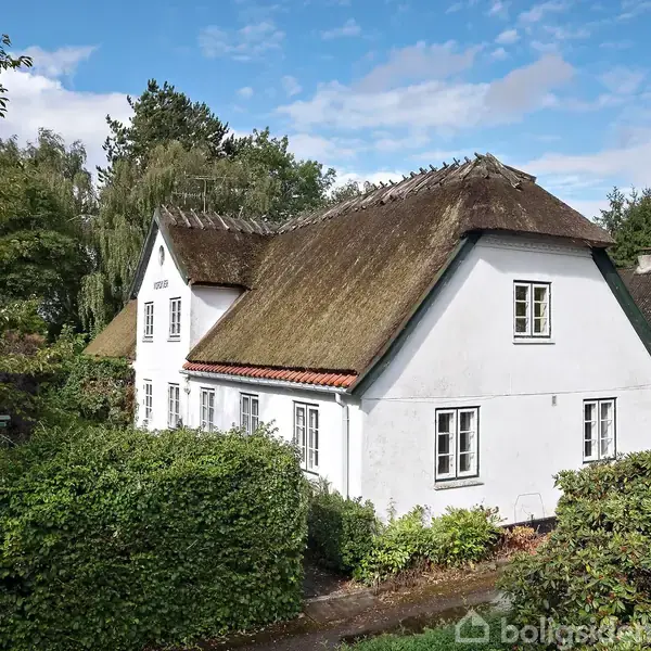 Et hvidt bindingsværkshus med stråtag står omkranset af tæt grøn beplantning. Omgivet af træer og en blå himmel skaber en idyllisk, landlig atmosfære.
