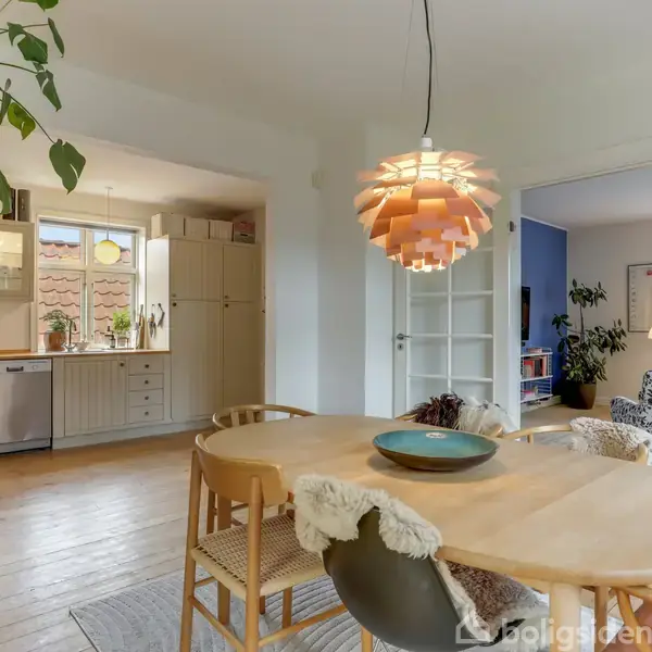 Et lyst spisebord fyldt med stole under en moderne lampe i et hyggeligt hjem. Til venstre er et åbent køkken med vindue, planter og en opvaskemaskine.