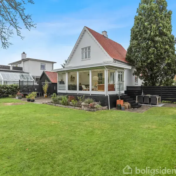 Et hvidt hus med en rød tag ligger i en have med græsplæne. En veranda ses foran huset, og der er et højt træ samt en hængekøje i baggrunden.