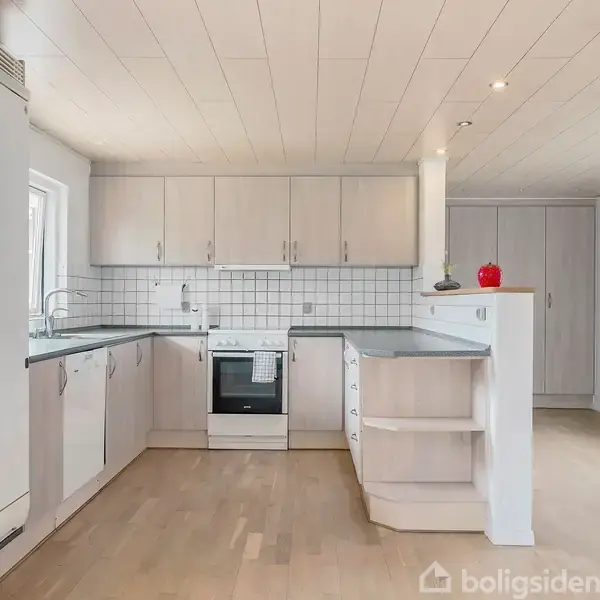 Et lyst køkken med hvide skabe og sort bordplade. En ovn er indbygget i midten, og der er en rød skål på disken som dekoration.