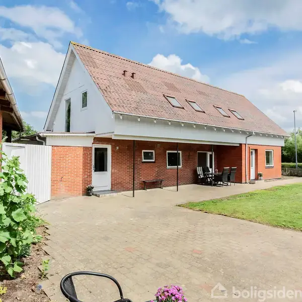 Rødt murstenshus med to-etagers tag, ventilerede vinduer og gårdhave. Terrassemøbler står placeret ved siden af huset, omgivet af græsplæne og blomsterbede under en blå himmel med skyer.