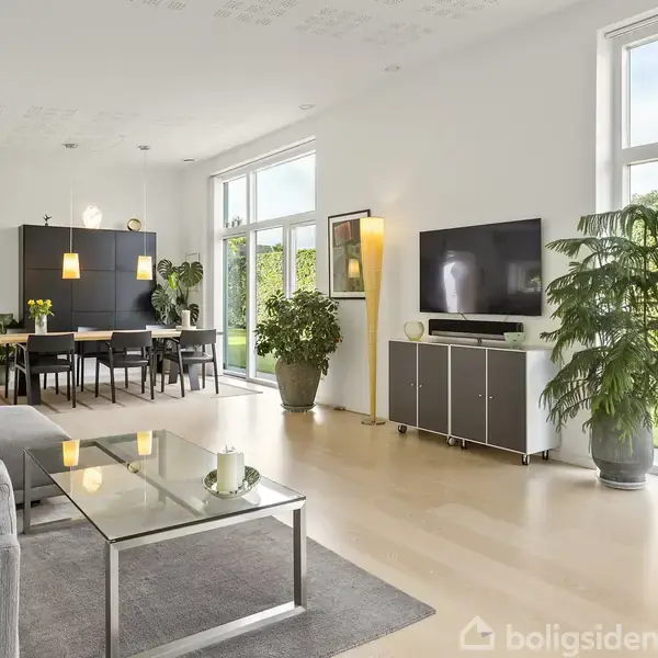 Stuen har en grå sofa, glasbord, og fjernsyn på skænk. Store vinduer giver udsigt til grøn hæk. Spisebord med stole er nær baggrund med loftlamper og sorte skabslåger.