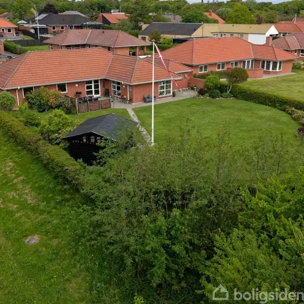 En rød murstensvilla med rødt tag og flagstang står i en frodig have omkranset af hække i et villakvarter med flere lignende huse.