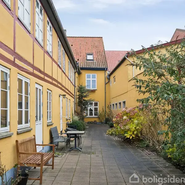 Gule bygninger med røde tage omgiver en brostensbelagt gårdsplads. En bænk og et bord står langs muren, mens buske vokser til højre. Himlen er overskyet.